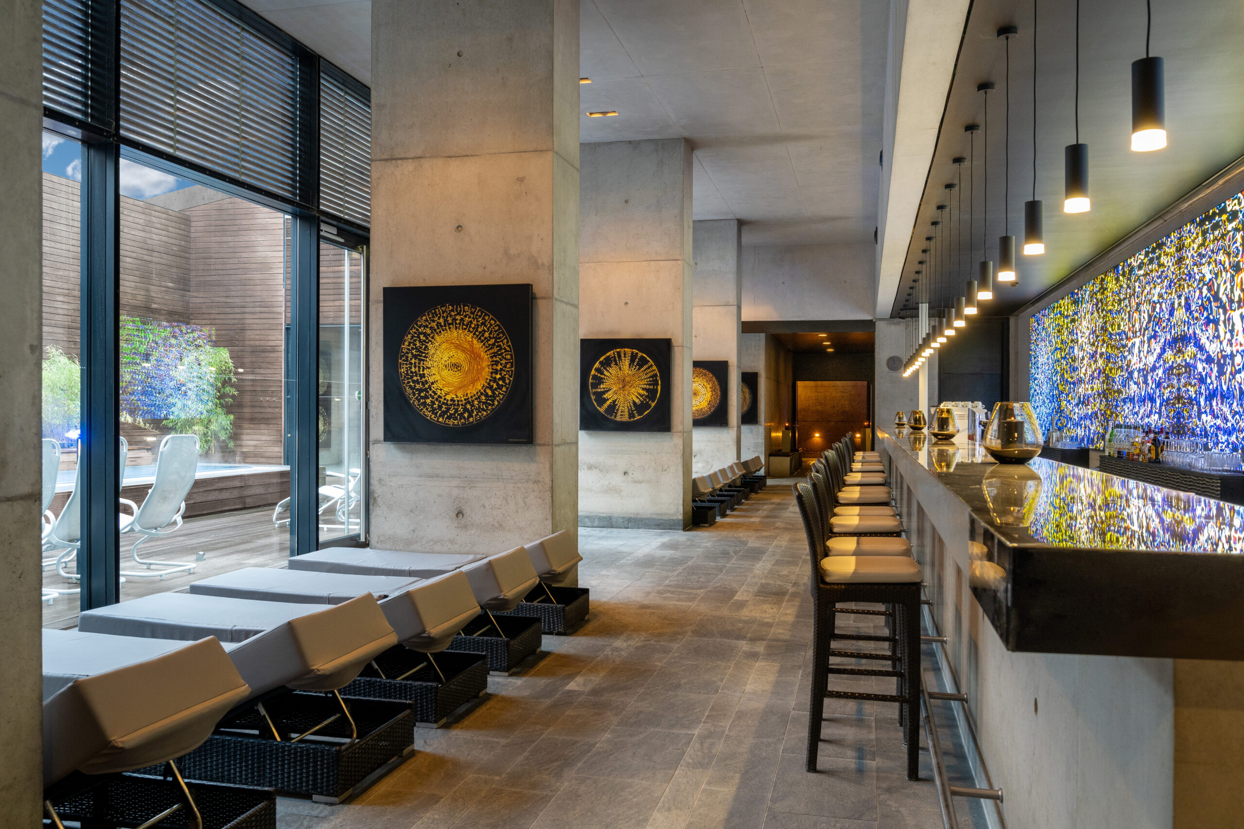 Eleganter Spa-Loungebereich mit Liegen am Fenster, modernen Kunstwerken an Betonwänden und stilvoll beleuchteter Bar mit leuchtendem Mosaik hinter dem Tresen.