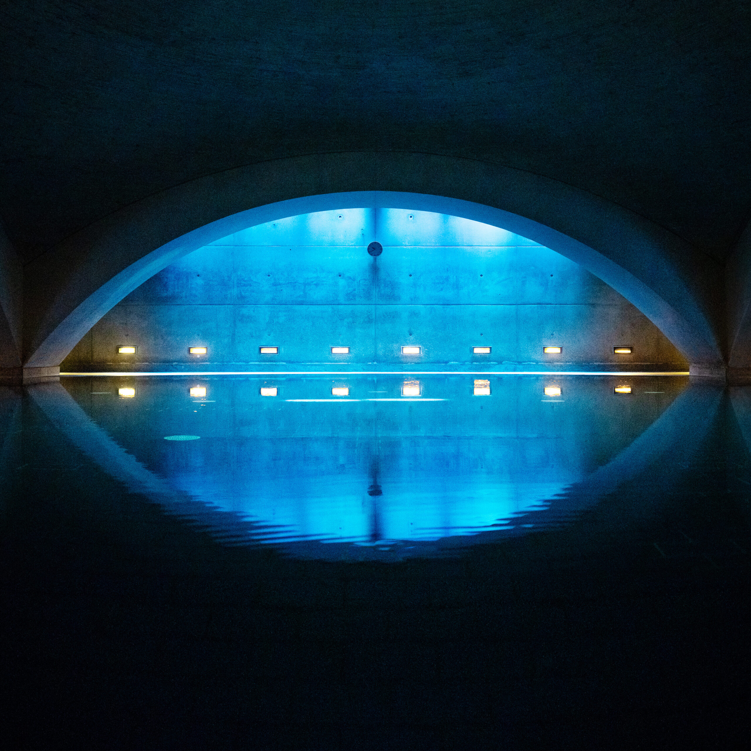 Frontale Ansicht eines ruhigen Hallenbeckens unter einem Gewölbe, mit blauer und gelber Akzentbeleuchtung, die sich auf der Wasseroberfläche spiegelt und eine meditative Stimmung erzeugt.