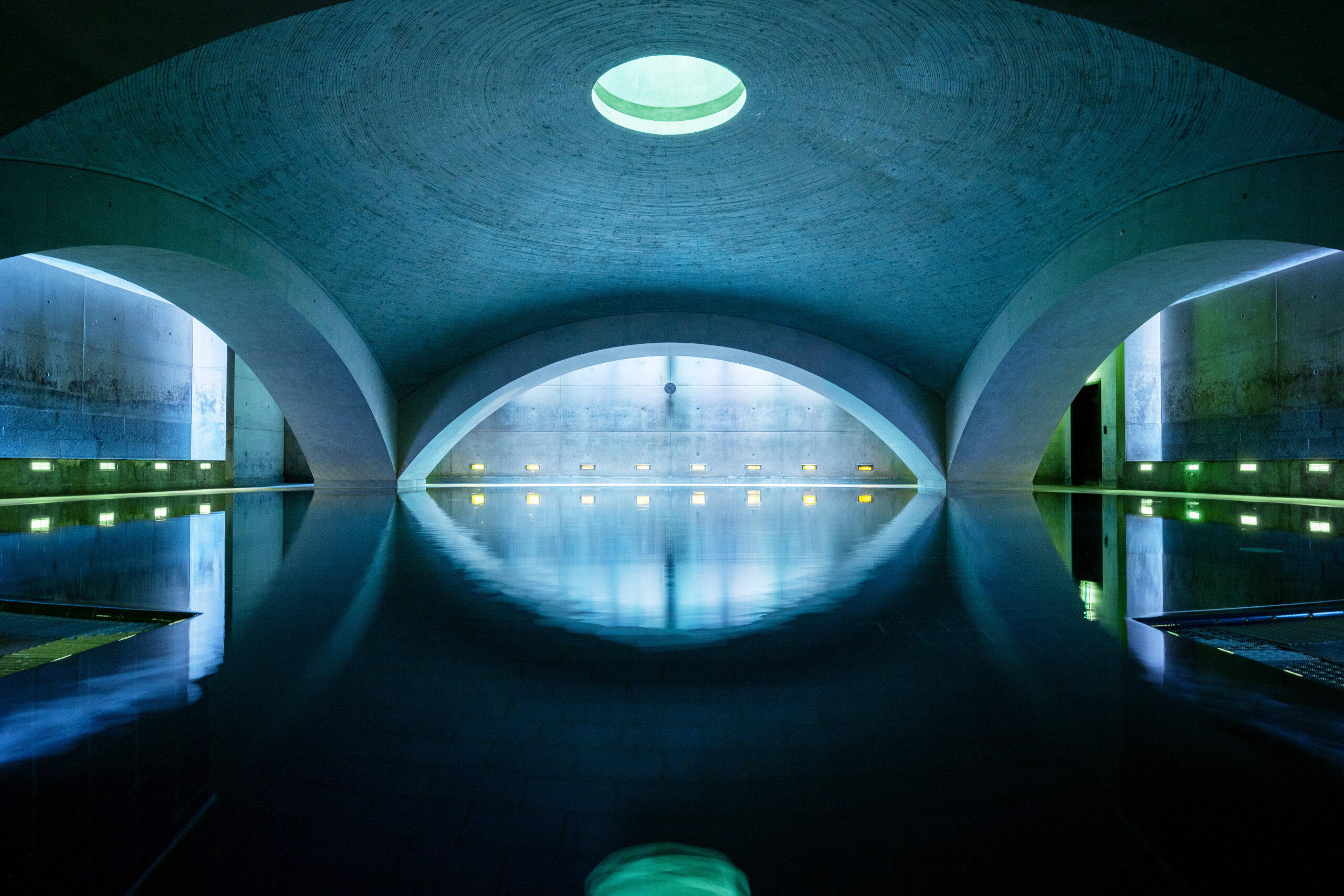 Weitwinkelansicht eines unterirdischen, gewölbten Betonraums mit zentralem Oberlicht, spiegelndem Wasserbecken und stimmungsvoller Wandbeleuchtung.