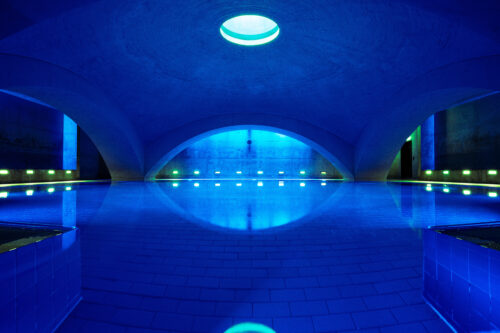 Weitwinkelaufnahme eines architektonisch markanten Hallenbads mit gewölbter Decke, intensiv blauer Beleuchtung, zentralem Oberlicht und stiller Wasseroberfläche mit gespiegeltem Lichtspiel.